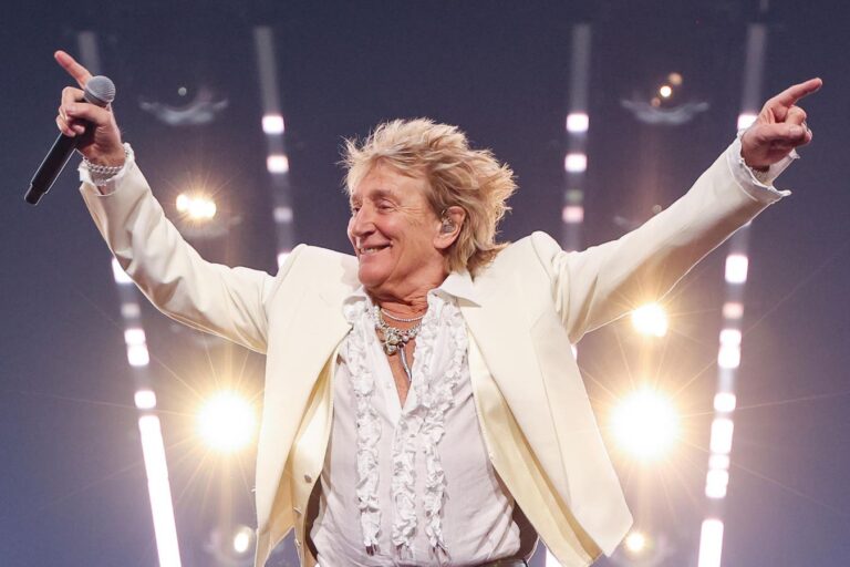 Rod Stewart se apresenta ao vivo usando terno branco e segurando um microfone