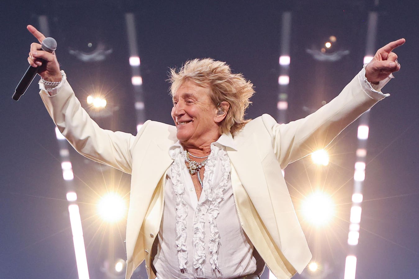Rod Stewart se apresenta ao vivo usando terno branco e segurando um microfone